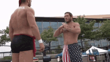 two men are standing next to each other in a wrestling ring . one of the men is wearing an american flag shorts .