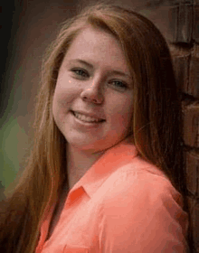 a young woman in an orange shirt is smiling and leaning against a brick wall .