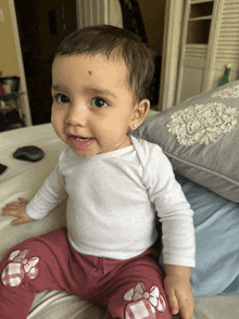 a baby with a red spot on her forehead sits on a bed next to a pillow