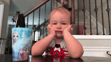 a little girl is sitting at a table with her hands on her face in front of a frozen trash can