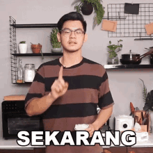 a man wearing glasses and a striped shirt is standing in a kitchen with the word sekarang written on the counter .