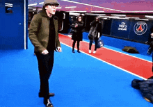 a man in a green jacket stands on a blue carpet in front of a wall that says paris saint-germain