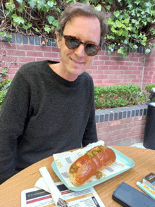 a man wearing sunglasses sits at a table with a hot dog on a tray that says lick