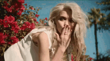 a woman is covering her mouth with her hand while standing in front of a bush of red flowers .