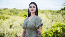 a woman in a green dress with a tattoo on her arm is standing in front of a forest .