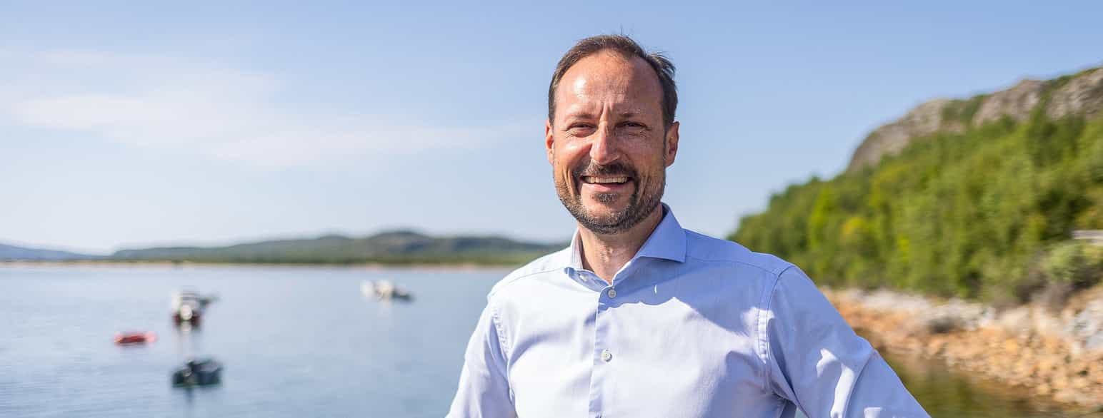 En mann i lys blå skjorte, med kortklipt mørkt hår og skjegg, smiler g myser litt mot kamera. Uskarp bakgrunn med noen små båter på vann, vannkant med ganske store steiner og noen grønne busker. Foto