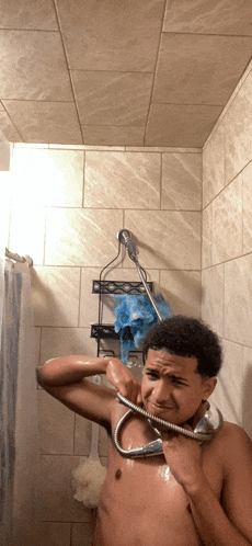 a shirtless man takes a shower with a shower head around his neck