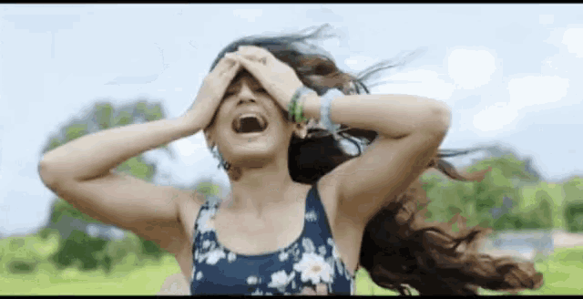 a woman in a floral tank top is covering her face with her hands while her hair blows in the wind