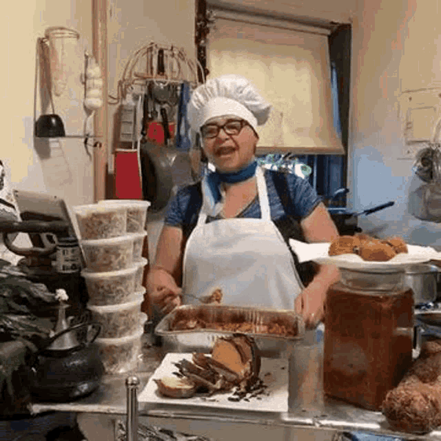 a woman wearing a chef 's hat and apron is standing in a kitchen holding a tray of food .