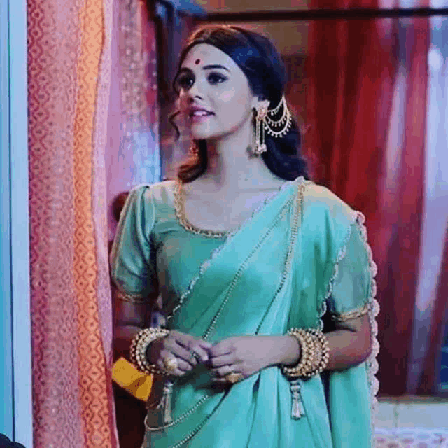 a woman in a green saree and gold jewelry is standing next to a window .