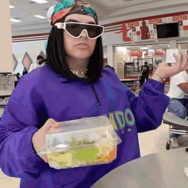 a woman wearing sunglasses and a purple sweater with the word vidor on it holds a plastic container of food