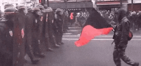 a man is walking down the street holding a red flag in front of a line of police officers .