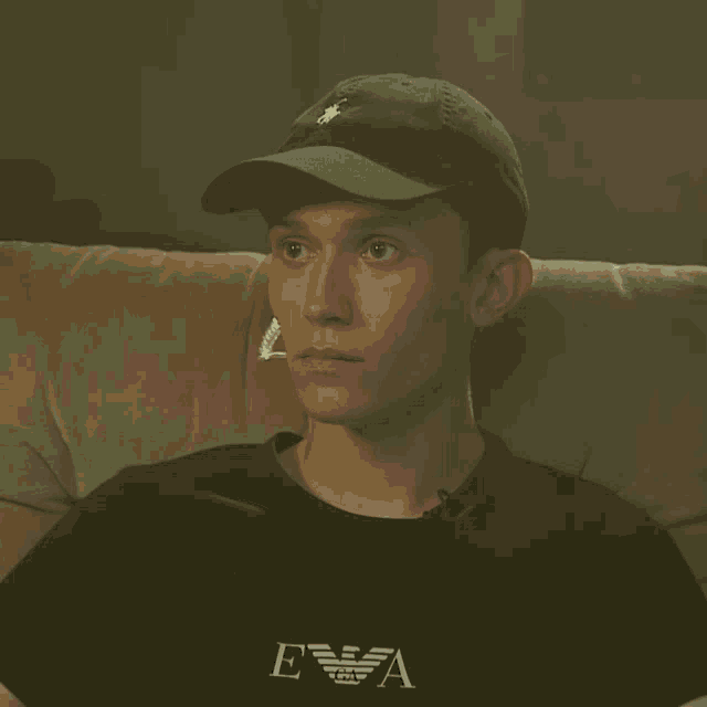 a young man wearing a hat and an emporio armani t-shirt sits on a couch