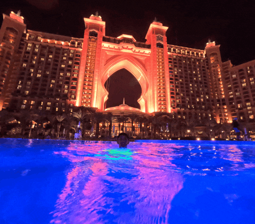 a man is swimming in a pool in front of a large building that is lit up at night