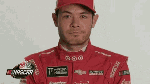 a man wearing a red racing suit and a red hat is standing in front of a nascar sign .