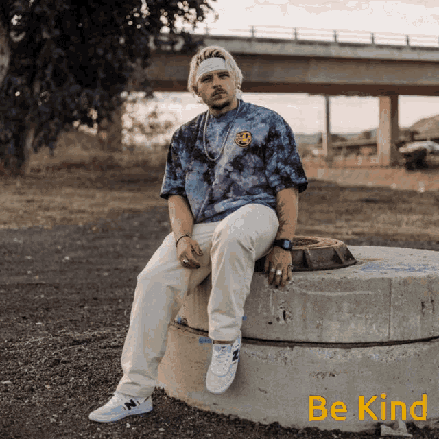 a man wearing a tie dye shirt sits on a concrete block with the words be kind written below him