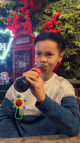 a young boy drinks from a coca cola bottle in front of a red phone booth