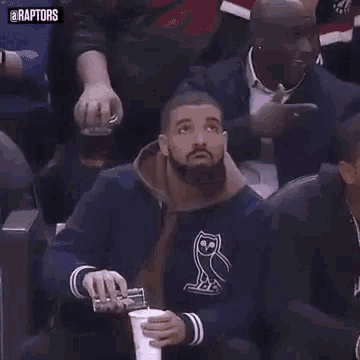 a man with a beard is holding a cup of coffee while sitting in the stands during a basketball game .