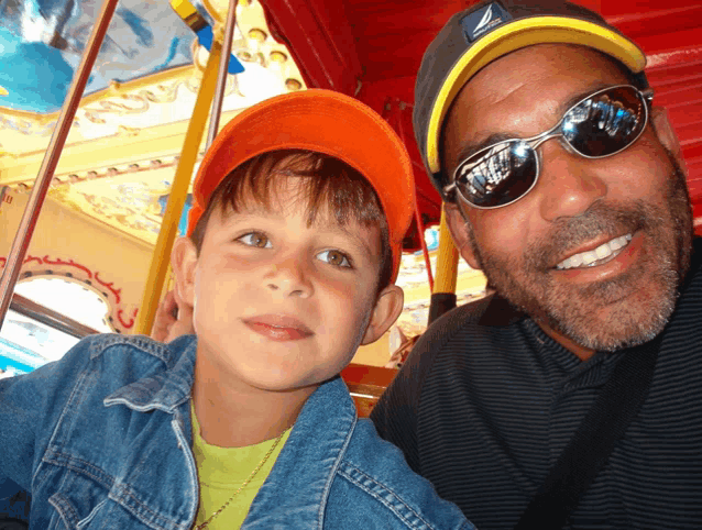 a man wearing sunglasses and a nautica hat smiles next to a boy wearing an orange hat