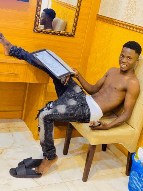 a shirtless man sits on a chair holding a laptop