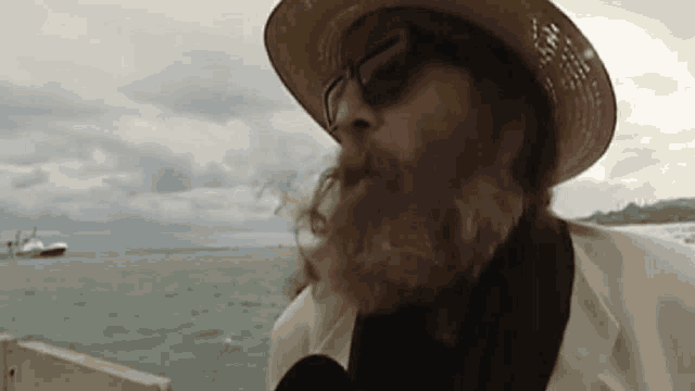 a man with a beard wearing a hat and sunglasses looks out over the ocean