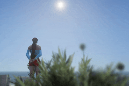 a woman in a blue shirt and red shorts stands in front of a blue sky with the sun shining brightly