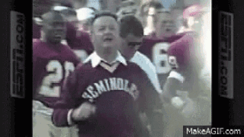 a group of football players are standing around a man wearing a sweater that says ' miami ' on it .