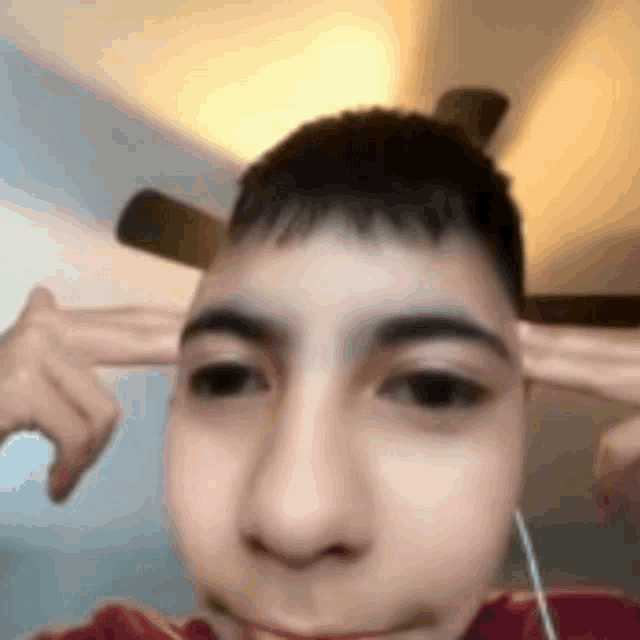 a young man is covering his ears with his hands while wearing headphones and a ceiling fan in the background .