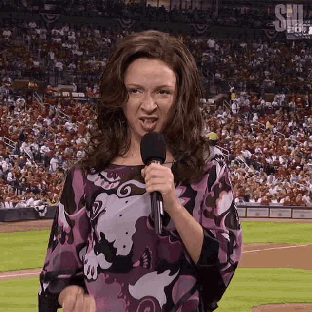 a woman in a purple shirt is holding a microphone in front of a crowd at a baseball game sponsored by fox