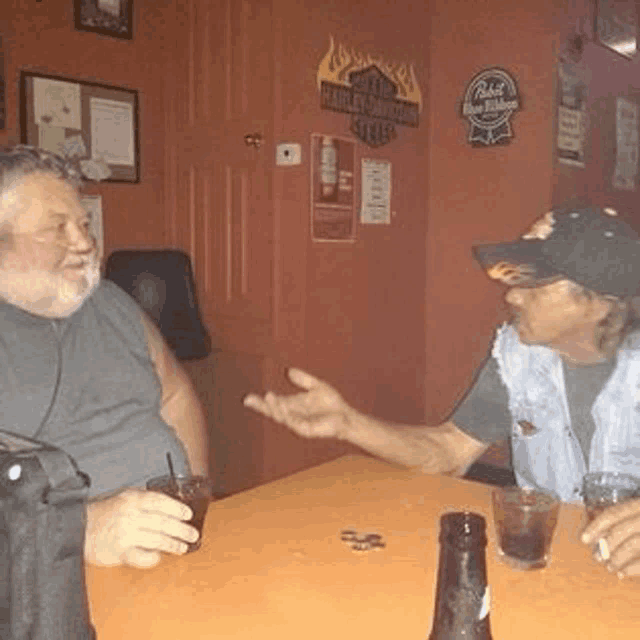 two men are sitting at a table talking to each other while holding drinks .