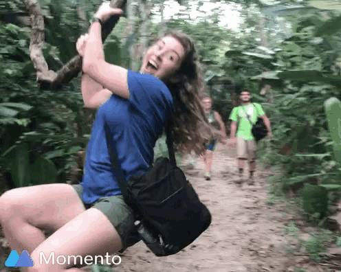 a woman in a blue shirt is swinging on a rope in the woods with a momento logo in the background