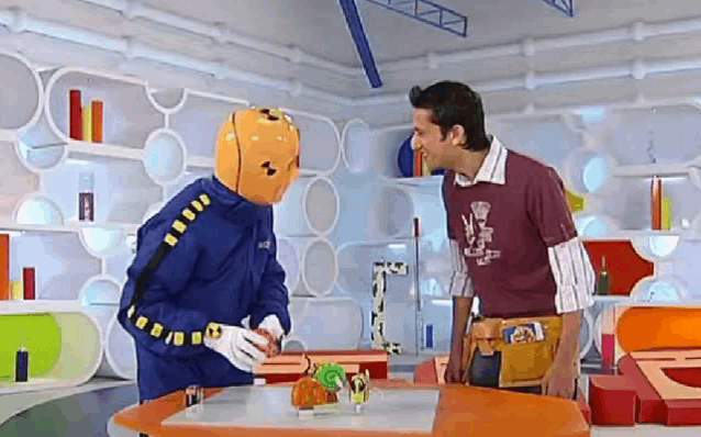 two men are standing next to each other at a table in a room . one of the men is wearing a yellow helmet .