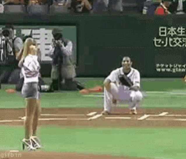 a woman is standing on a baseball field in front of a man kneeling on the field .