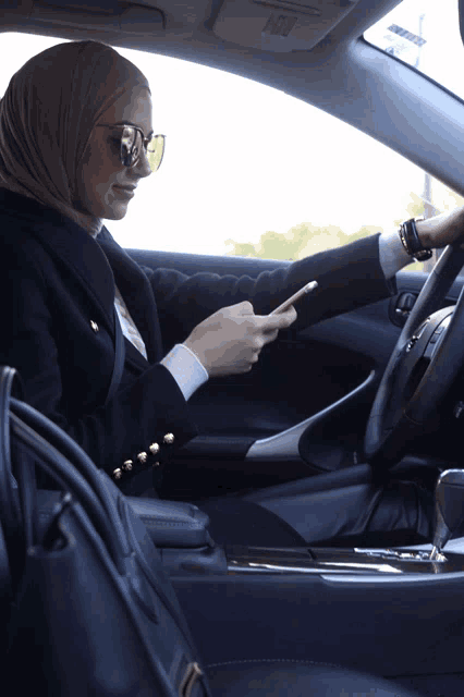 a woman in a hijab is sitting in a car looking at her cell phone