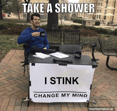 a man is sitting at a table with a sign that says take a shower i stink change my mind