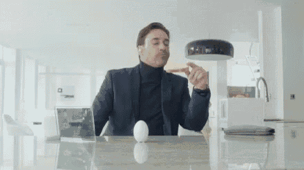 a man in a suit and turtleneck is sitting at a table with an egg .