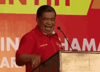 a man in a red shirt is laughing while standing at a podium with the word ham on the wall behind him