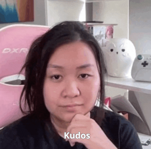 a woman is sitting in front of a pink chair with her hand on her chin and the word kudos written on her face .