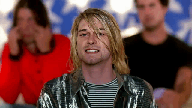 a man with long blonde hair and a striped shirt smiles for the camera