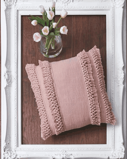 a pink pillow with fringe sits on a wooden floor next to a vase of tulips