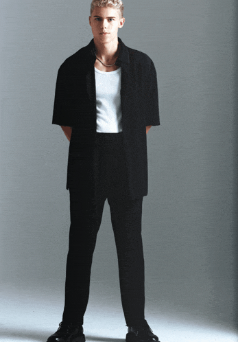 a man wearing a black shirt and black pants stands in front of a white wall