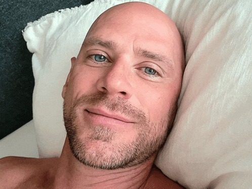 a bald man with a beard and blue eyes is laying on a bed