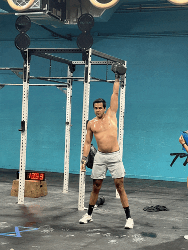 a shirtless man is lifting a dumbbell in a gym with a digital clock displaying 13:53