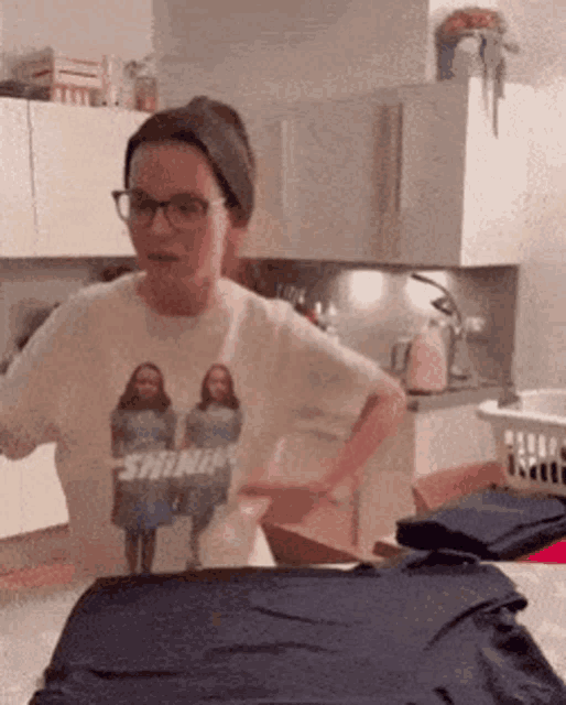 a woman wearing glasses and a t-shirt with a picture of two girls on it is standing in a kitchen .