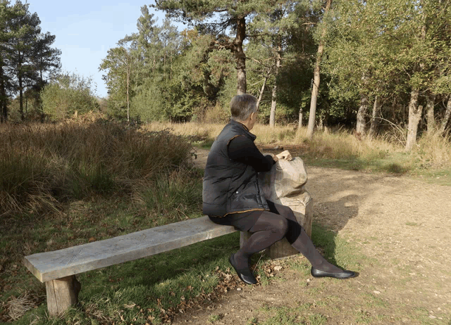 a woman sits on a bench in the woods holding a bag