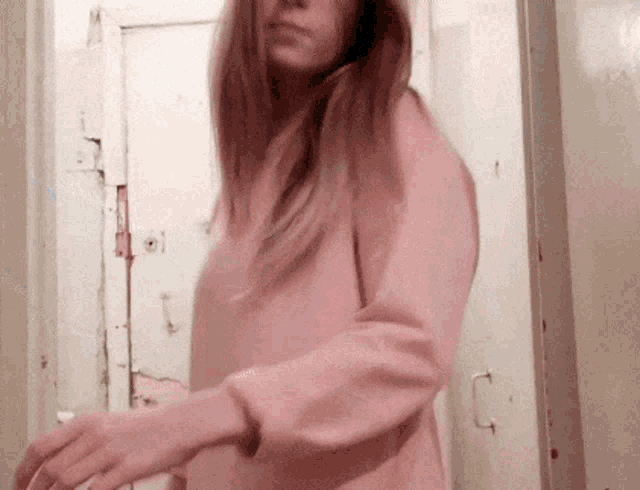 a woman in a pink sweater is standing in front of a door and looking at the camera .