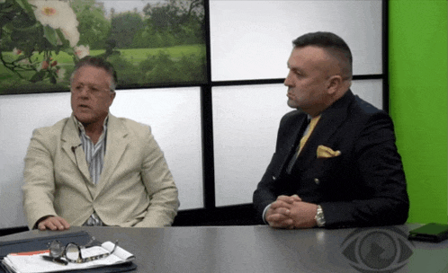 two men are sitting at a table with their hands folded in front of a green wall