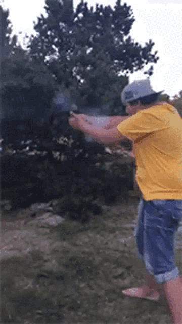 a man in a yellow shirt is holding a gun in front of a tree