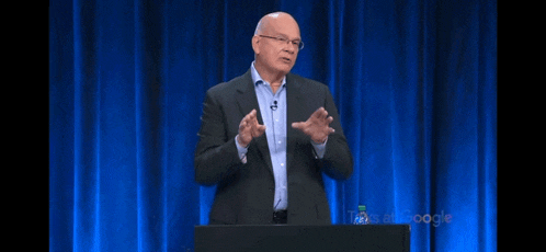 a man in a suit is giving a speech on a stage with a blue curtain behind him that says google