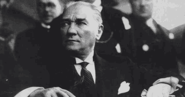 a black and white photo of a man in a suit and tie sitting in front of a crowd .